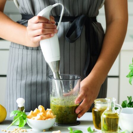 A cropped image of a person in front of a kitchen using a hand blender to make a green puree. The kitchen counter has multiple cooking ingredients on it